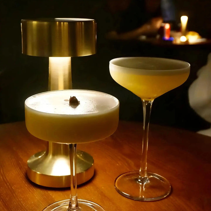 Two elegant cocktails in stemmed glassware with frothy tops, illuminated by a stylish table lamp.